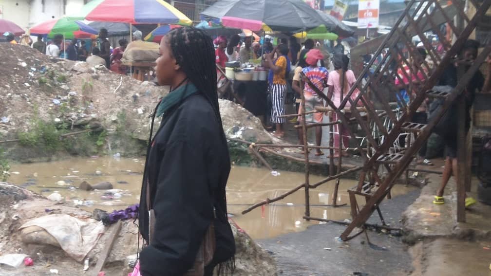 At Tombo-Waterloo junction in Waterloo town, a lady stood by beside a created hole by workers of WAPP. ©️Africa24/photo credit: Hassan I. Conteh